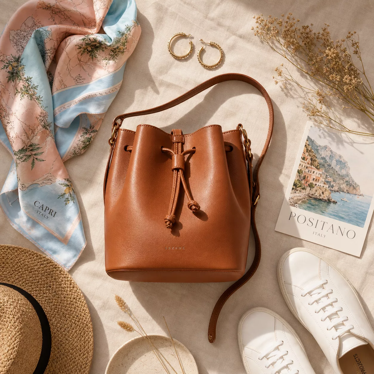 A stylish flat lay arrangement on a light linen surface featuring a caramel brown leather bucket bag as the centerpiece, surrounded by summer fashion accessories including a delicate silk scarf in pastel pink and sky blue tones, minimalist white sneakers, a pair of gold hoop earrings, and a few sprigs of dried flowers. Warm natural sunlight filters across the scene creating soft shadows. The overall aesthetic is clean, modern, and summery with a Mediterranean mood. Shot from above in editorial fashion photography style with a warm, airy color palette of cream, caramel, sage green, and blush tones.