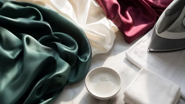 A beautifully composed flat-lay photograph showcasing flowing silk fabric in rich, luxurious colors — deep emerald green, ivory white, and burgundy red — elegantly draped and layered on a clean marble surface. Beside the fabric, a small white ceramic bowl contains a few drops of gentle liquid soap, and a soft white cotton cloth is neatly folded nearby. A clothes iron rests at the edge of the frame on its lowest heat setting, with a thin protective white cloth beneath it. Warm, soft natural light falls across the scene from the side, highlighting the luminous sheen and delicate texture of the silk. The overall mood is calm, refined, and editorial — evoking luxury care, craftsmanship, and the artful preservation of fine textile. Realistic photography style, high detail, shallow depth of field.