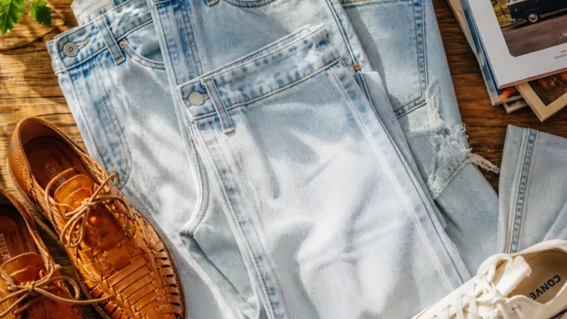 A beautifully styled flat-lay editorial photograph featuring light-washed ice denim jeans in shades ranging from pale sky blue to near-white, arranged artfully on a warm natural wooden surface bathed in soft diffused sunlight. Nearby, a pair of honey-toned handcrafted leather shoes and white canvas sneakers are placed as complementary accessories. The composition evokes a breezy, effortless spring aesthetic with a nostalgic yet modern feel. Warm natural light casts gentle shadows across the fabric, highlighting the characteristic faded wash texture of the denim weave. The overall mood is relaxed, confident, and casually stylish, with a vibrant editorial quality and rich, saturated colors.