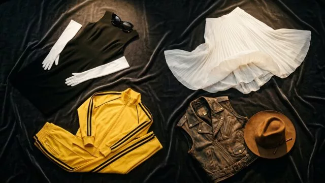 A cinematic flat-lay composition featuring iconic fashion pieces arranged dramatically on a dark velvet surface: a sleek little black dress with long white gloves and oversized sunglasses, a flowing white pleated skirt catching an imaginary breeze, a vibrant yellow tracksuit with black racing stripes, and a worn leather vest beside a brown wide-brimmed fedora hat. Soft golden spotlight illuminates each piece from above, casting elegant shadows. The overall mood is glamorous, editorial, and nostalgic, evoking the golden age of Hollywood costume design. Rich textures, deep shadows, and a sophisticated color palette of black, white, yellow, and warm brown. Photorealistic studio photography style with a cinematic, high-fashion editorial aesthetic.