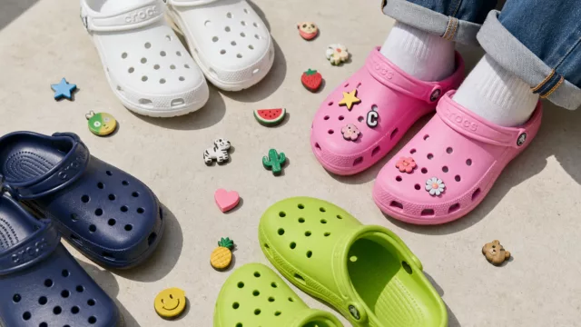 A flat-lay styled photograph of colorful original Crocs clogs in multiple vibrant colors — including classic white, bubblegum pink, navy blue, and lime green — arranged artfully on a clean light concrete surface. Several small Jibbitz charms are scattered around the shoes, including star, fruit, and animal shapes. One pair is styled with a rolled denim jean cuff visible, suggesting a casual streetwear aesthetic. Soft natural daylight illuminates the scene from the side, creating gentle shadows. The composition is editorial and modern, with a fashion-forward yet approachable mood, shot from a slightly elevated 45-degree angle.